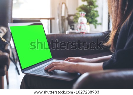 Mockup image of woman using and typing on laptop with blank green desktop screen while sitting on sofa
