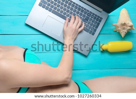 Remote work on the laptop in a seaside resort. A girl in a swimsuit is using a laptop. Accessories for relaxation on the beach: sunglasses, sunblock, headphones, shell on a blue wooden background. 