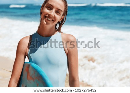 Satisfied happy female surfer being glad after going in for sport, stands wet against ocean view with surfing board, smiles happily at camera. Joyful sporty woman in bathing suit. Active rest concept