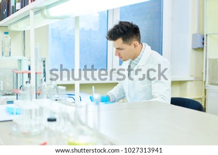 Profile view of handsome young researcher wearing rubber gloves and white coat sitting at desk and taking notes while carrying out experiment in modern lab