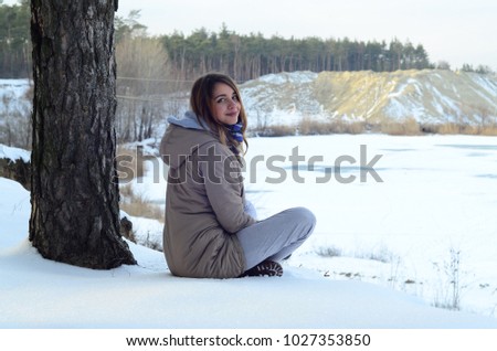 A young Caucasian girl in a brown coat is sitting near a cliff in the background of a horizon line between the sky and a frozen lake in winter time