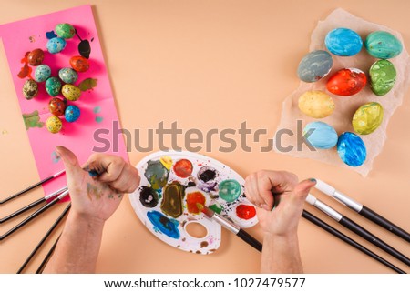 Top view of colored chicken and quail easter eggs, brushes, palette, paints and dirty hands showing thumb up gesture on beige pastel background. Egg as an Easter symbol. Creative process.