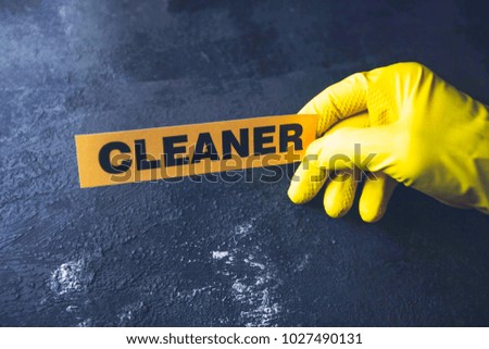 Cleaning office or house concept. Mens hand in yellow rubber glove holding Cleaner inscription sign, concrete dark background, top view, copy space