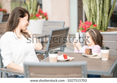 Cute little girl taking a picture of her mother with mobile phone while sitting at a restaurant