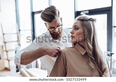 Handsome male fashion designer is taking measurements from attractive female model while working in his workshop. Stylish bearded man in process of creating new clothes collection.