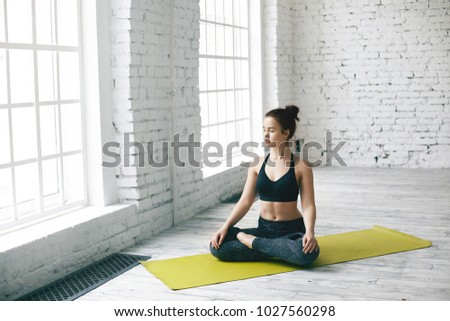 Isolated shot of beautiful brunette young female wearing sports outfit practicing yoga on fitness mat, closing eyes, keeping legs crossed in lotus pose, having calm peaceful look on her face