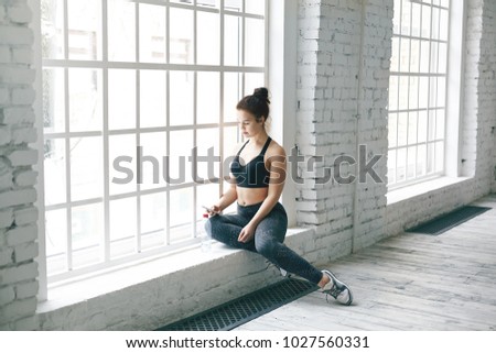 Picture of attractive young woman fitness instructor sitting on windowsill in spacious room before group workout, searching for music tracks on her mobile phone. People, sports and technology