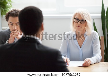 Serious attentive senior female hr manager employer listening to candidate at job interview, focused strict mature businesswoman thinking about hiring decision at difficult group negotiations concept Royalty-Free Stock Photo #1027563259