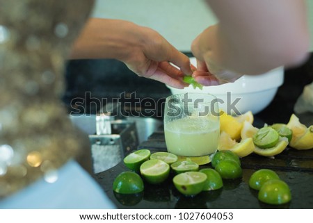 Asian lady hands squeeze green lime juice in clear glass with many pieces of fresh lime on black table.