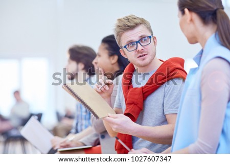 Young man in eyeglasses pointing at notepad while looking at colleague and asking her question