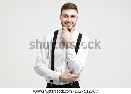 Style and fashion concept. Isolated picture of positive young unshaven male entrepreneur of Caucasian appearance wearing elegant formal clothes smiling cheerfully, touching his well trimmed beard