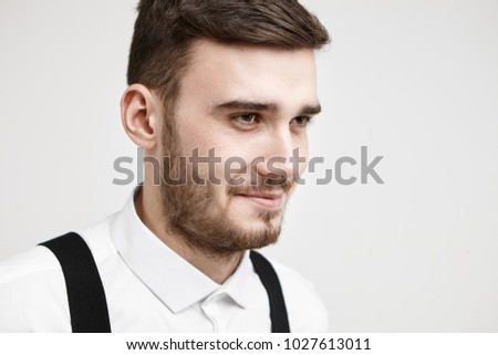 Half profile picture of friendly looking fashionable young male with mustache and beard smiling thoughtfully as he reminds some funny story or joke, posing in studio wearing white formal shirt