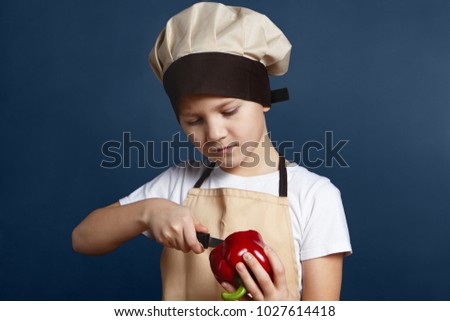 Health, nutrition and food. Picture of serious concentrated little boy in chef cap standing at blank wall and peeling red pepper with knife while cooking healthy dinner or lunch with fresh vegetables