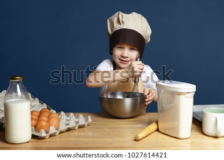 Picture of little boy wearing apron and chef cap whisking ingredients in metal bowl while cooking pancakes, cookies or other pastry, standing at kitchen table with eggs, milk, flour and rolling pin