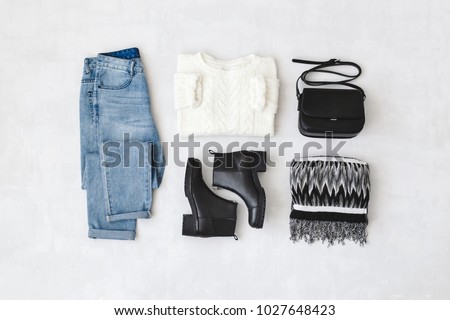 Blue jeans, white knitted sweater, small black cross body bag, leather ankle boots and striped scarf on grey background. Overhead view of woman's casual day outfits. Trendy hipster look. Flat lay. Royalty-Free Stock Photo #1027648423