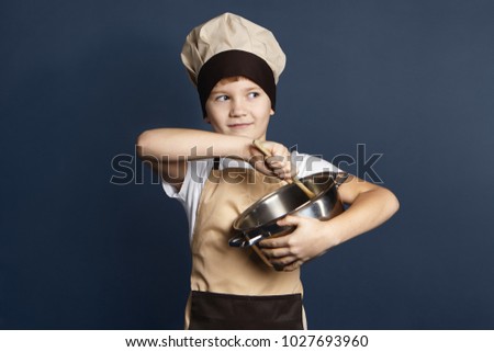 Isolated picture of cute European teen boy in apron and hat cooking something in studio, holding casserole and looking sideways with mysterious smile, preparing surprise dinner for his parents.