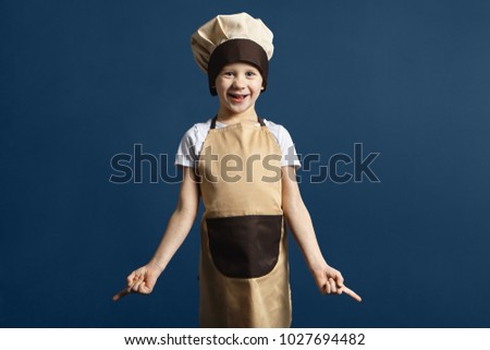 Isolated picture of emotional funny Caucasian little boy in chef uniform having excited look, opening mouth and pointing fingers down. Human facial expressions, emotions, feelings and reaction