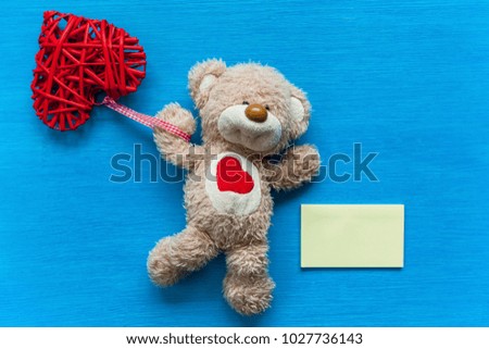 Beige, teddy bear lies on a turquoise table and holds a heart. Bear with a heart.