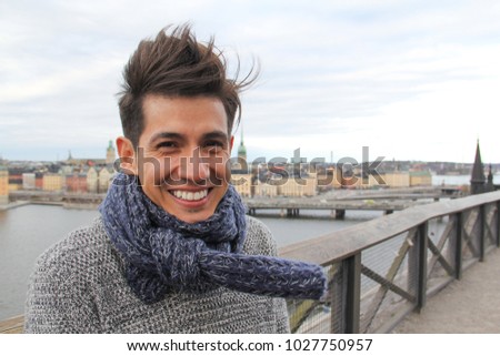 A handsome young Latino man braves the cold and wind, with warm blue scarf, and hair blown by wind, with view of European city in background.  Looking direct to camera. Room for text / copy. Royalty-Free Stock Photo #1027750957