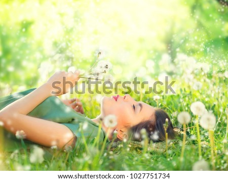 Beautiful Young Woman lying on the field in green grass and blowing dandelion. Outdoors. Enjoy Nature. Healthy Smiling Girl on spring lawn. Allergy free concept. Freedom