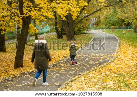 Mother and daughter are walking in the autumn in the park. Little girl is riding a scooter
