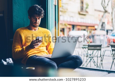 Bearded graphic designer reading incoming notification on smartphone connected to 4G internet while working freelance at computer.Hipster guy checking message on cellular during studying at netbook