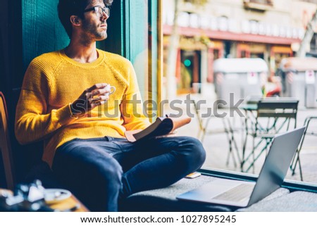 Thoughtful young man in eyeglasses looking out of window and thinking on new creative startup project while holding cup of tasty coffee in hand sitting with laptop in coffee shop. Crative blogger