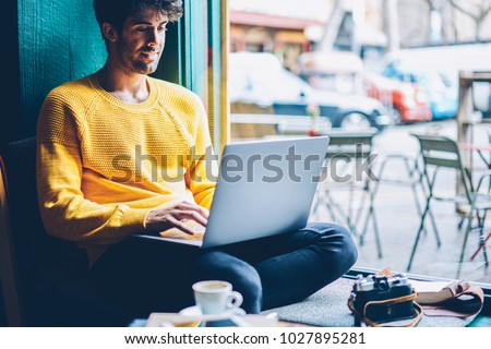 Smiling hipster guy chatting online with friends during leisure time at laptop device connected to wireless 4G internet.Cheerful male blogger publishing post on website on netbook resting in coworking Royalty-Free Stock Photo #1027895281