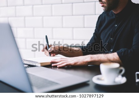 Cropped view of stylish businessman dressed in black shirt making notes in notepad working at modern laptop computer in coffee shop interior. Student writing in notebook sitting at netbook device