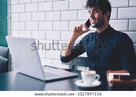 Successful entrepreneur having mobile conversation with operator manager on smartphone device while working at laptop computer connected to wireless internet sitting in stylish coffee shop interior
