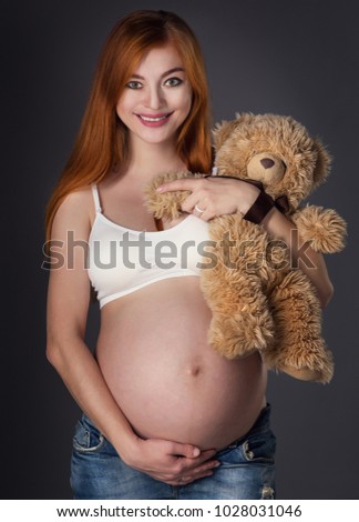 Beautiful red-haired pregnant girl in top and jeans on a gray background with a toy bear in hands