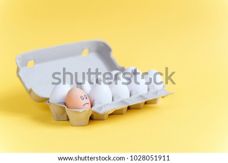 One orange egg with drawn sad face among white eggs in a cardboard tray on yellow background. Different from other.