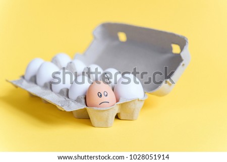 One orange egg with drawn sad face among white eggs in a cardboard tray on yellow background. Different from other.