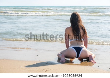 Unrecognizable woman bruenette with long hairs in a bikini sits on the sand beach. Back view.