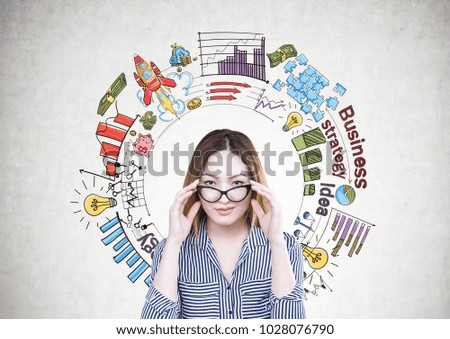 Attractive and serious Asian businesswoman wearing a striped shirt is touching her glasses and looking at the viewer. A concrete wall background with a business plan sketch