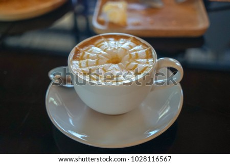 Caffe latte with latte art design in spider web flower pattern on milky foam layer in white cup and saucer with light reflection on black glass table and blurred background, Thailand