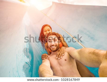 Happy crazy couple taking selfie photo with action camera in aqua park - Young people having fun in summer holidays with technology trends - Vacation, youth, love and travel concept - Focus on faces 