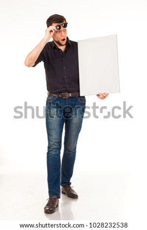 Stylish man posing with a white sign in his hands (for posting advertising, logo). Dressed in a black shirt and jeans and black glasses. Isolated on white background