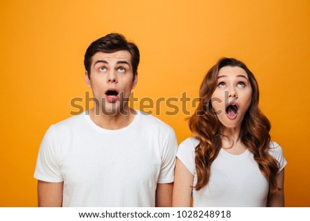 Photo of excited boyfriend and girlfriend in white t-shirts looking on camera with open mouth isolated over yellow background