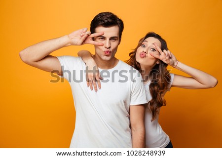 Image of happy lovely friends smiling and posing on camera while showing victory signs together over yellow background