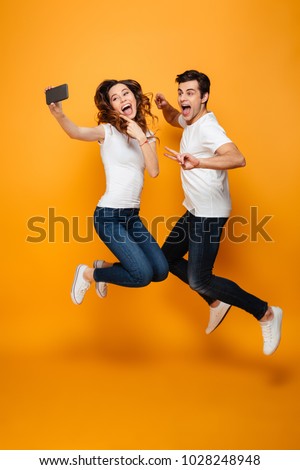 Image of two joyful man and woman 20s making selfie on cell phone while jumping and gesturing together, over yellow background
