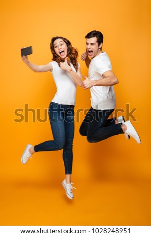 Portrait of two happy people man and woman making selfie on smartphone while jumping together, over yellow background