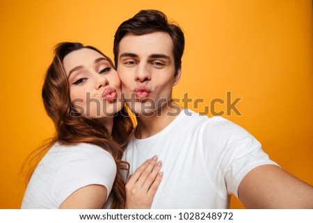 Portrait of two joyful male and female people making selfie while doing kissing lips together on camera over yellow background