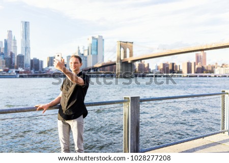Young man outside outdoors in NYC New York City Brooklyn Bridge Park by east river, railing, looking at view of cityscape skyline, taking selfie with smart phone