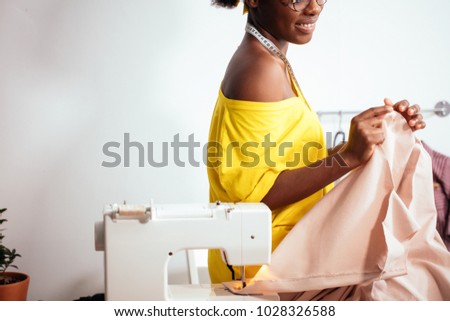 Young african woman seamstress looking at pink fabrics and standing in workshop