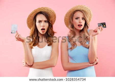 Portrait of a two shocked girls dressed in swimsuits and summer hats holding credit cards isolated over pink background