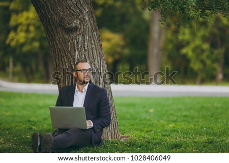 Young successful businessman in white shirt, classic suit, glasses. Man sit on grass ground, work on laptop pc computer in city park on green lawn outdoors on nature. Mobile Office, business concept