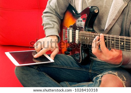 Casual young man in ripped jeans, playing sunburst offset electric guitar. Young musician taking online musical instrument course lesson on wireless internet, tablet gadget. Background, close up.