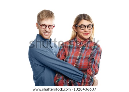 People, relationships, love, romance and dating concept. Picture of beautiful blonde woman having disgusted look while being held tight by weird unshaven nerdy male against white wall background
