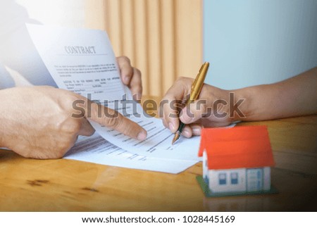 Close up of customer signing a paper document for buying house. Estate agent pointing finger showing where to sign on wooden desk: real estate, home loan and insurance concept